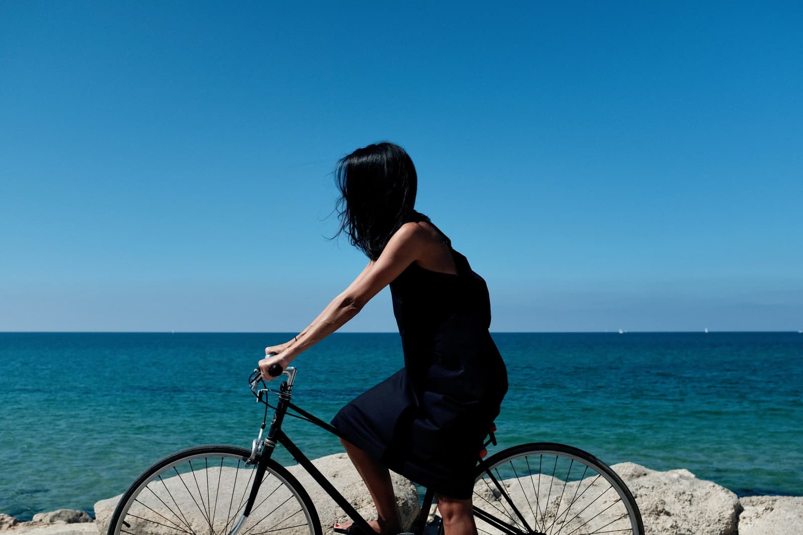 Cycliste en robe noire sur un vélo de ville, face à une mer turquoise